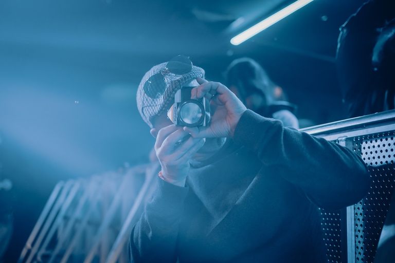 Photographer Daniel Down wearing a cap and holding a vintage Fujica camera to his eye under cool blue stage lights at a concert venue.